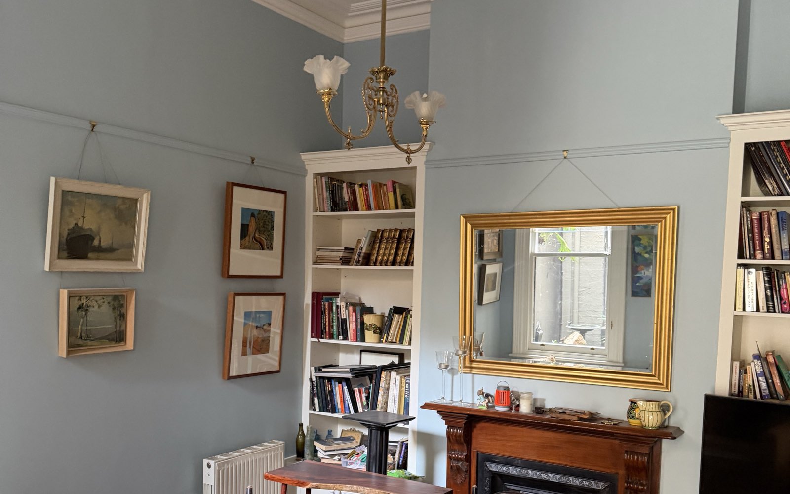A period living room painted in blue-grey, with a moulded cornice, chandelier, built-in bookshelves, a gilt overmantel mirror and a timber fireplace surround.